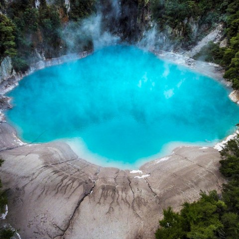 Vibrant blue geothermal pool surrounded by dense green forest and rocky terrain.