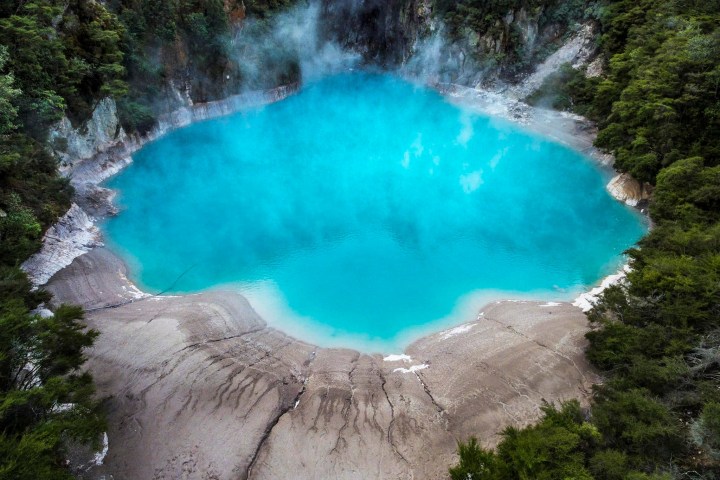 Vibrant blue geothermal pool surrounded by dense green forest and rocky terrain.