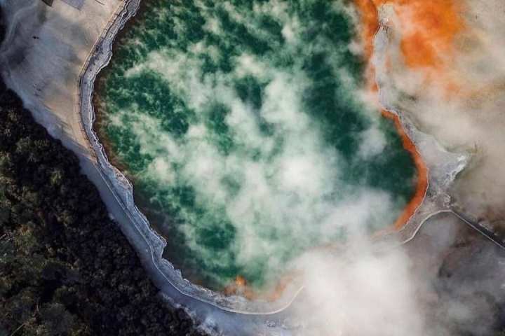 Aerial view of a steaming hot spring with green water and orange edges.
