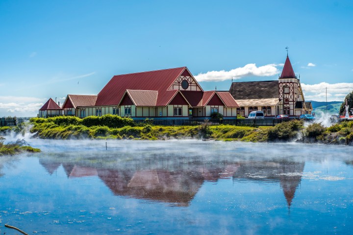 Buildings with red roofs and reflections in a steaming lake under a blue sky.