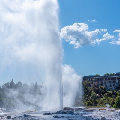 Geyser erupting with steam under blue sky; trees and building in background.