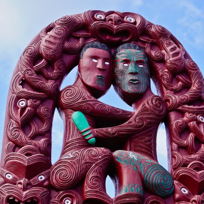 Detailed wood carving of two figures embracing, with ornate patterns and a blue sky background.