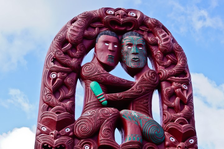Detailed wood carving of two figures embracing, with ornate patterns and a blue sky background.