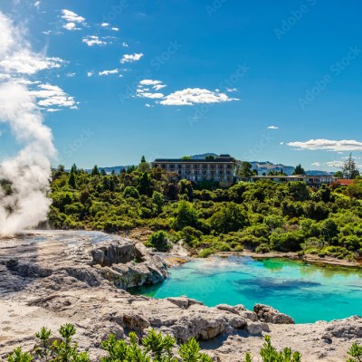 Geyser emitting steam near a turquoise pool with lush greenery and a building in the background under a blue sky.