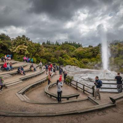 Tourists gather around a geyser with wooden seating and trees under a cloudy sky.