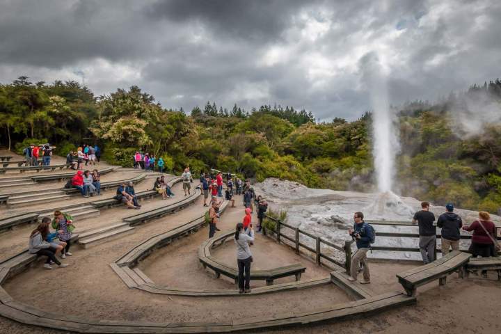 Tourists gather around a geyser with wooden seating and trees under a cloudy sky.