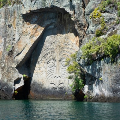 Rock face carving with intricate patterns and greenery at water's edge.