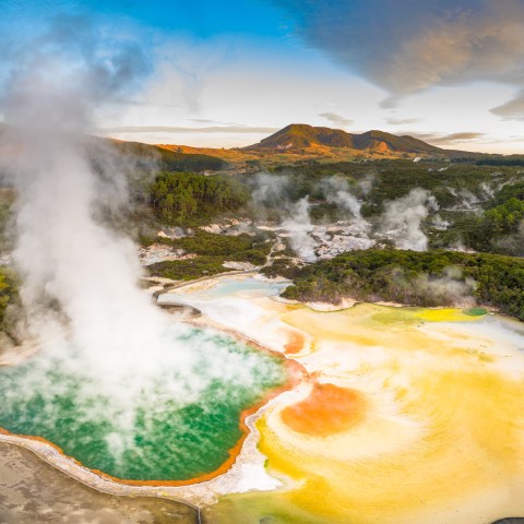 Aerial view of geothermal hot spring with colorful pools emitting steam, surrounded by lush forest and hills.