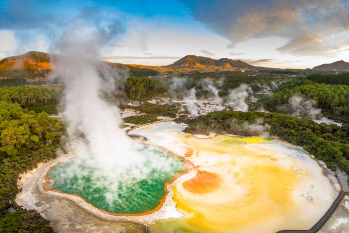 Aerial view of geothermal hot spring with colorful pools emitting steam, surrounded by lush forest and hills.