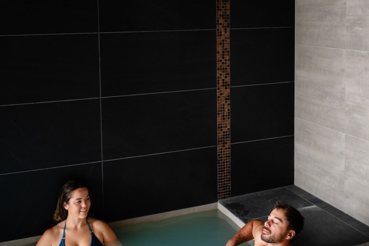 Two people relaxing in an indoor spa pool, with dark and light tiled walls.